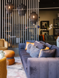 Modern living room with a blue sofa, decorative pillows, leather ottomans, and hanging lights behind a slatted divider.