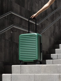A bottle green hard shell Samsonite carry on wheel bag, sitting on grey stairs.