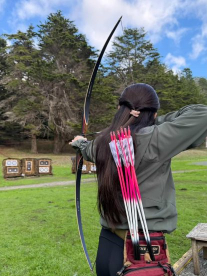 Person aiming a bow and arrow at outdoor archery targets on a grassy field, viewed from behind.